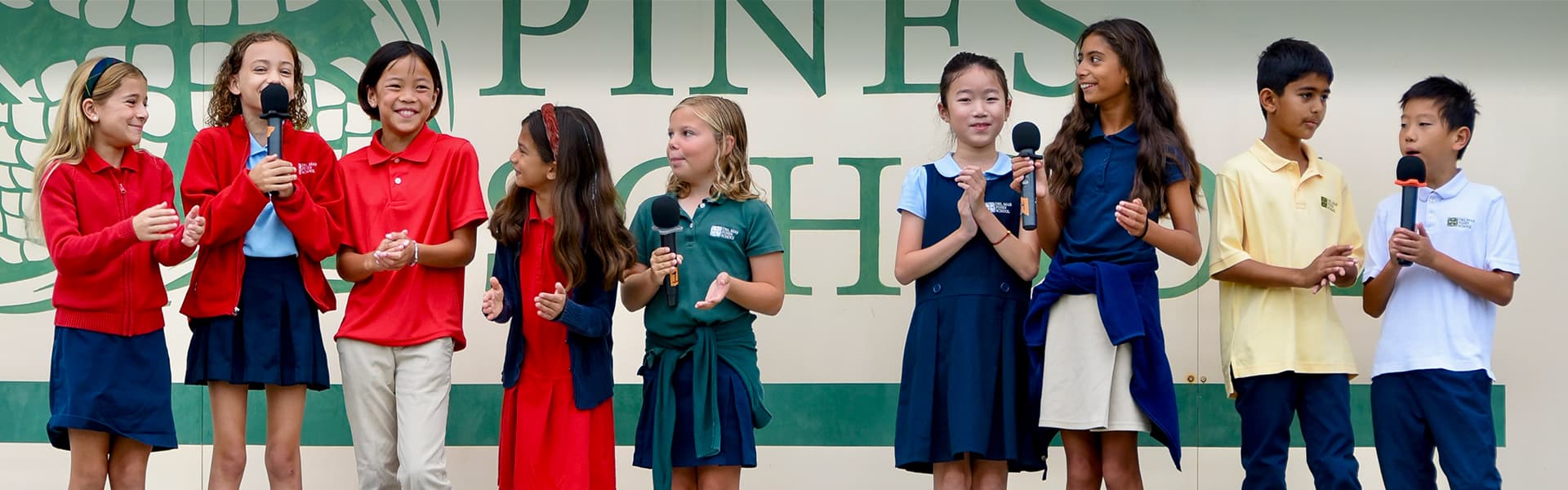 Students on stage clapping and holding microphones in front of the Pines School mural