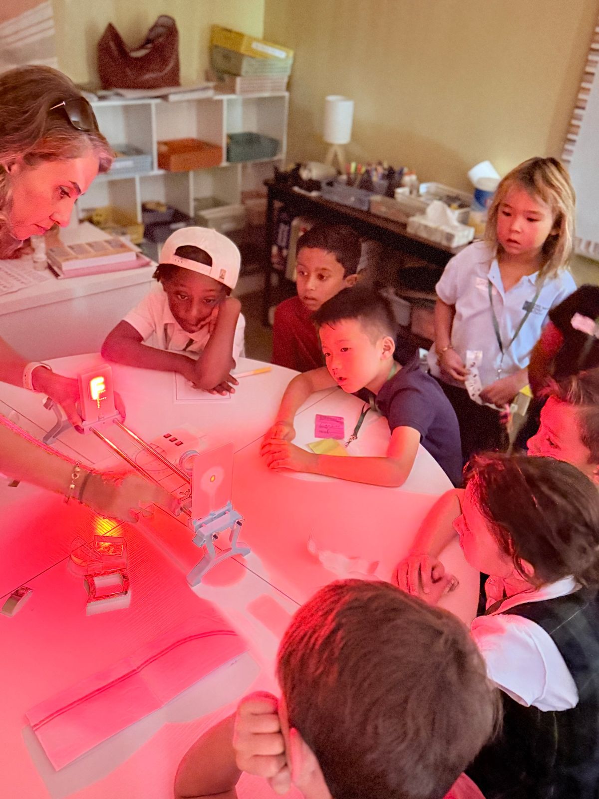 Students gathered around a table glowing with red light while a parent presenter demonstrates a light experiment.