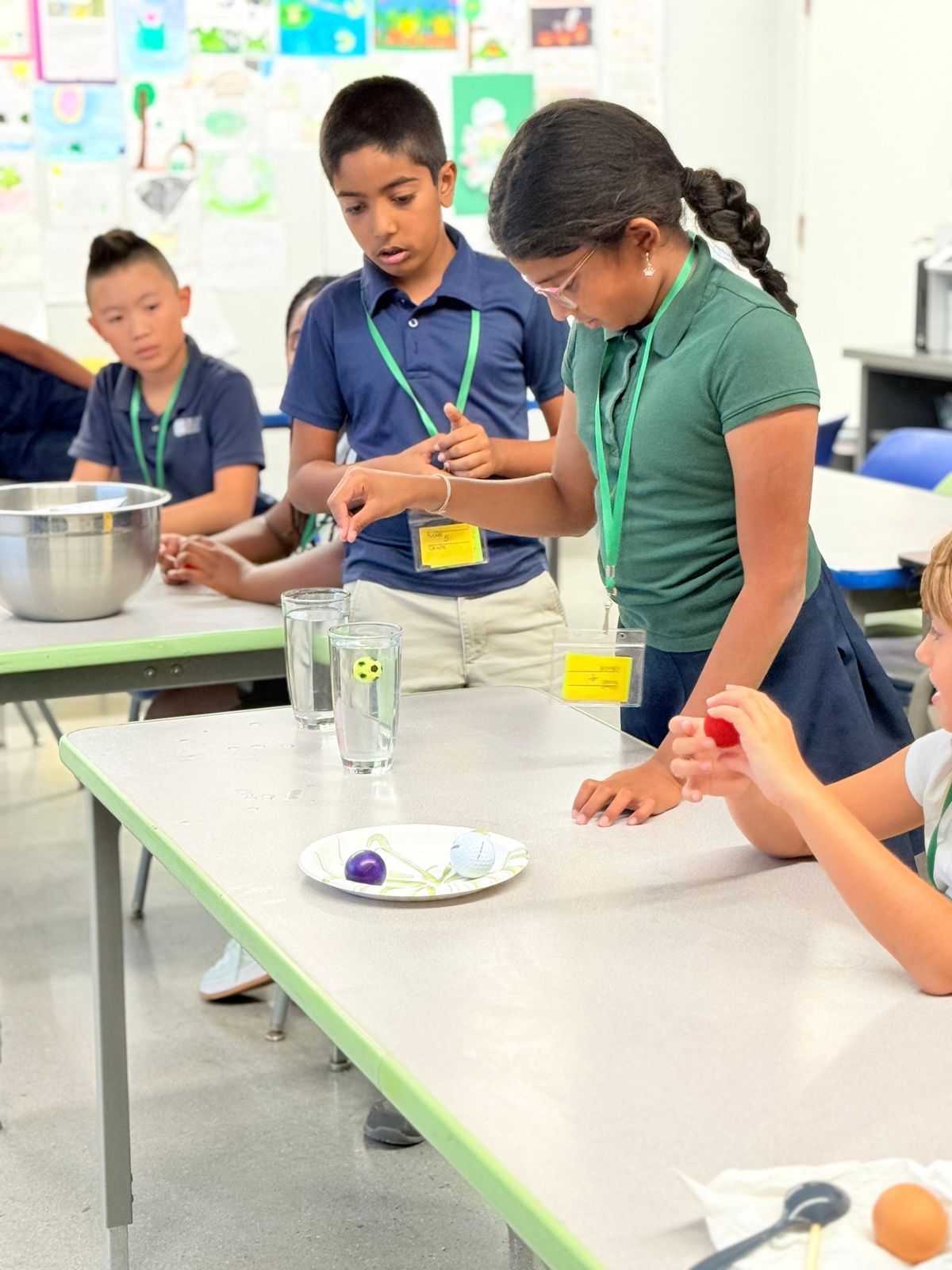 Students at a classroom table watching a floating-ball experiment in glasses of water.