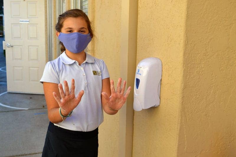 A DMP student wearing a face mask and school uniform stands outside by a wall-mounted hand sanitizer dispenser, holding up her hands covered with sanitizer and smiling at the camera.