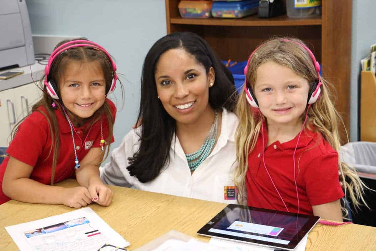 A teacher sits at a table with two young girls wearing headphones. The children are smiling, wearing red shirts, and there is a tablet and papers on the table. Shelves with supplies are in the background.