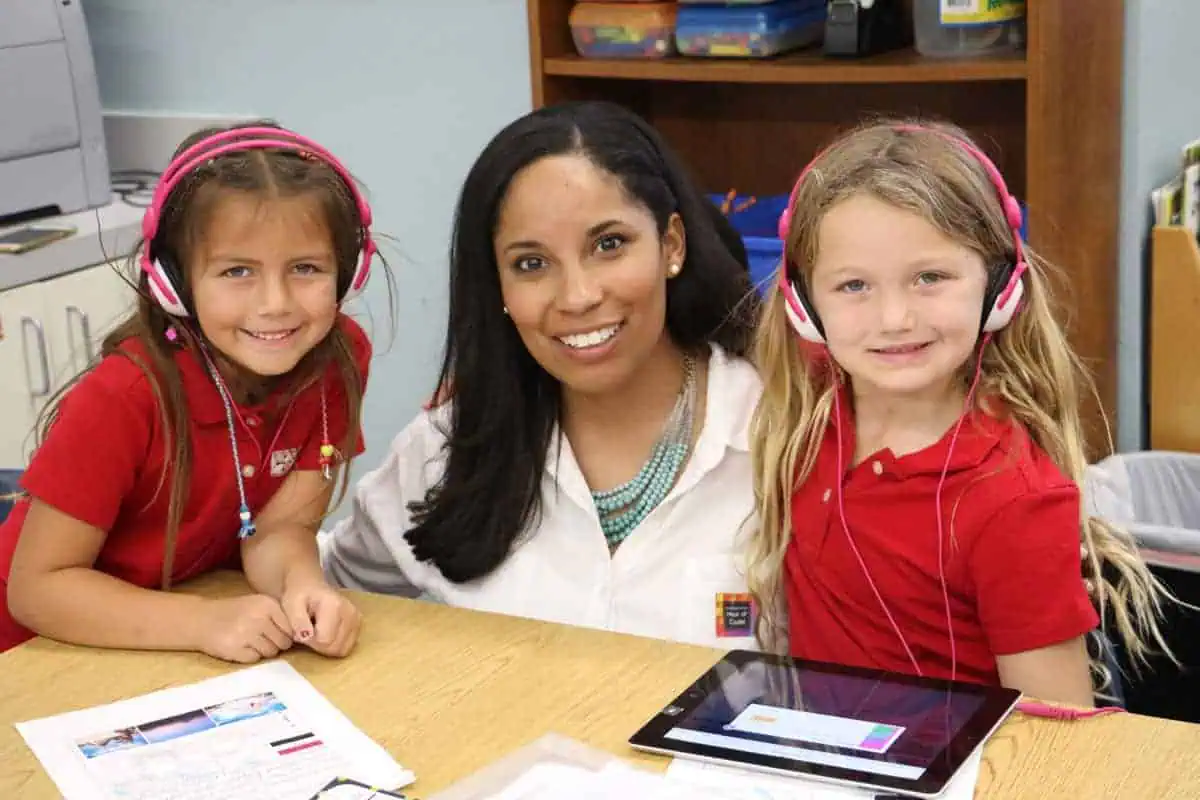 A teacher sits at a table with two young girls wearing headphones. The children are smiling, wearing red shirts, and there is a tablet and papers on the table. Shelves with supplies are in the background.