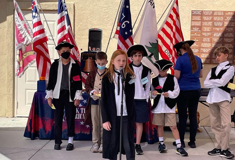 DMP students dressed in colonial-era costumes stand in front of American flags. A girl speaks into a microphone while others stand behind her,.