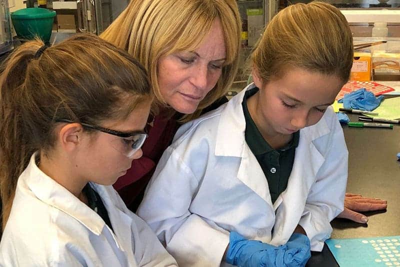 A DMP teacher closely observes and guides two girls wearing lab coats and gloves as they work on a science experiment in a laboratory setting.
