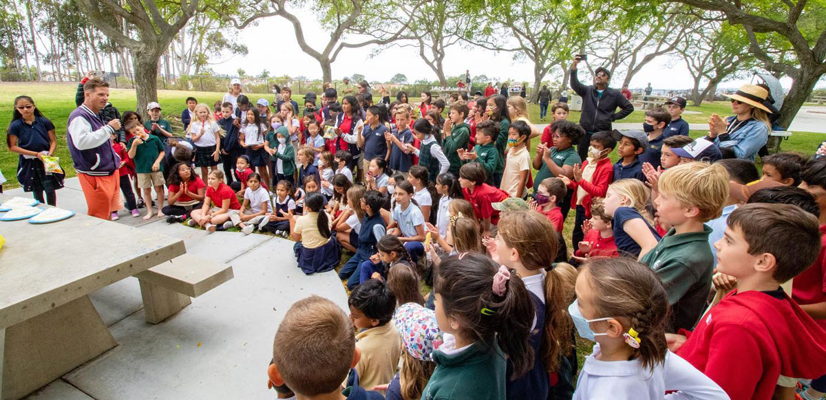 Del Mar Pines students sit and stand outdoors in a park, attentively watching an adult speaking near a picnic table, surrounded by trees on a sunny day.