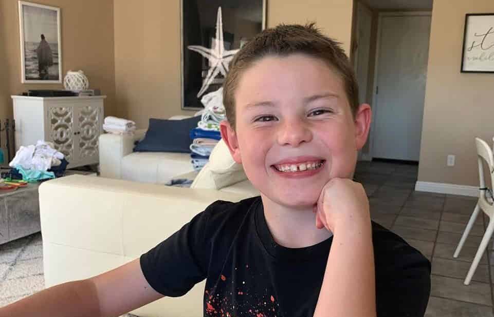 A young boy with short brown hair smiles at the camera, resting his chin on his hand.