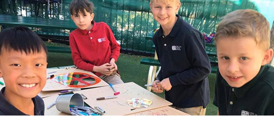 Four young boys smile and work on art projects at an outdoor table, surrounded by markers and colorful drawings.