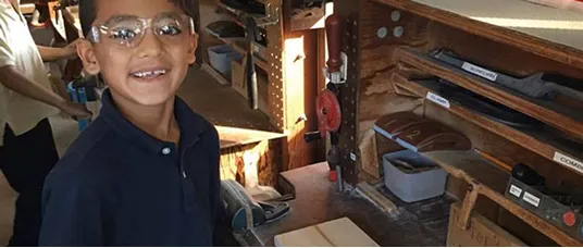 A young student wearing safety glasses and a navy shirt smiles while standing at a woodworking bench with various tools and materials organized on shelves in the background.