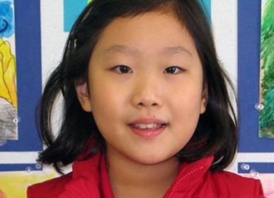 A young girl with short black hair, wearing a red jacket, smiles softly at the camera. Colorful artwork is displayed on the wall behind her.