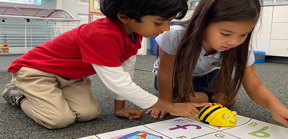 Two Del Mar Pines students kneel on the floor, guiding a bee-shaped robot across a colorful alphabet mat in a classroom setting.