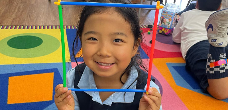 A young girl smiles while holding a square frame made of colorful sticks in front of her face, sitting on a bright, geometric-patterned rug in a classroom.