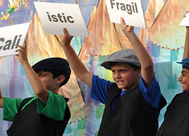 Three children wearing hats and vests hold up signs in front of a colorful, painted backdrop.