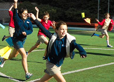 Children run and laugh on a grassy field while flying colorful kites or paper airplanes.
