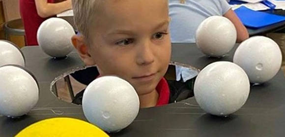 A young boy dressed as the solar system, with a circular black board around his neck featuring white spheres representing planets and a large yellow sphere symbolizing the sun.