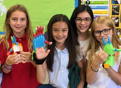 Four smiling girls stand together in a classroom, each holding colorful 3D-printed prosthetic hands.