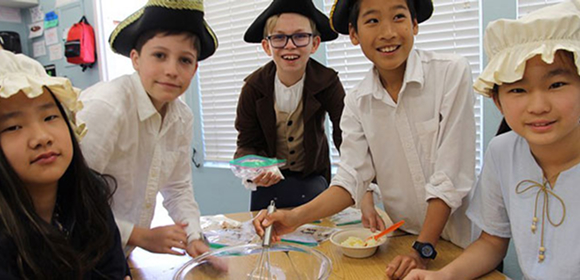 Five students dressed in historical costumes smile and work together around a table, mixing ingredients in bowls, likely participating in a classroom activity or reenactment.