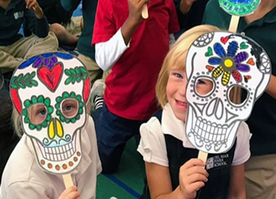 Two young children sit on the floor, holding up colorful Day of the Dead skull masks in front of their faces, surrounded by classmates in a classroom setting.