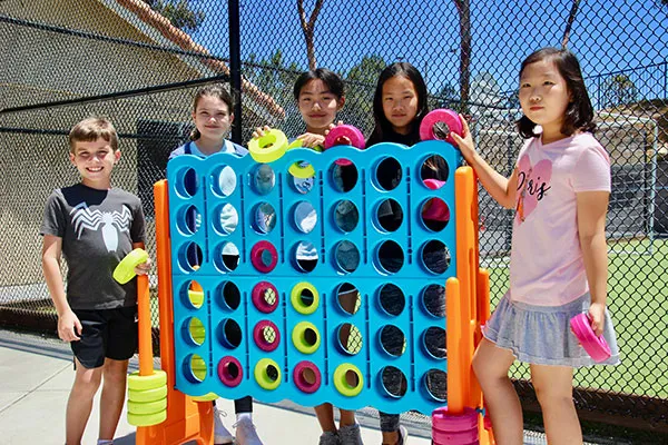 Five children stand outdoors next to an oversized Connect Four game, smiling and holding game pieces.