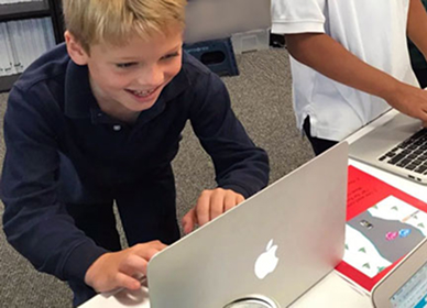 Two girls smiling and sitting together in a classroom, with other students and colorful artwork visible in the background.