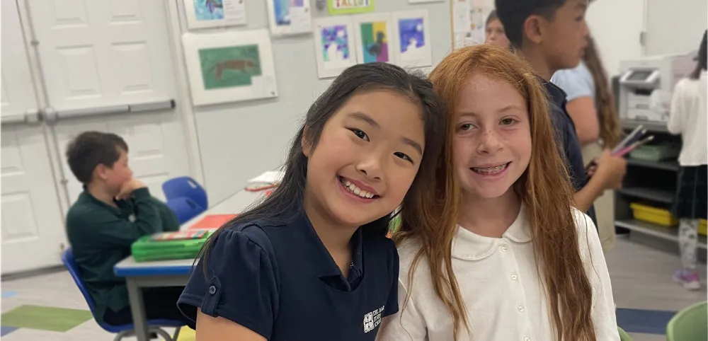 A young girl wearing a black face mask stands in a classroom, holding up a Eureka Math Grade 4 Modules 1 & 2 book. Classroom decorations and cabinets are visible in the background.